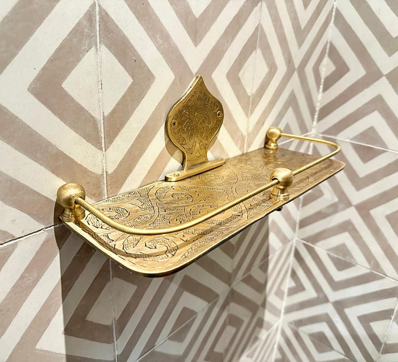 Puede incluir: Un estante dorado montado en la pared con un dise&ntilde;o ornamentado y grabado. El estante tiene un frente curvo con un borde elevado y una placa trasera decorativa. Una barra de metal curvada recorre el borde frontal. El estante est&aacute; montado en una pared de azulejos estampados.