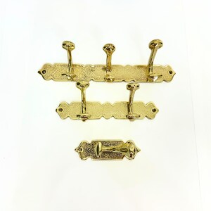 Pode incluir: Conjunto de tr&ecirc;s cabides dourados e um gancho individual. Cada cabide tem uma superf&iacute;cie texturizada e bordas decorativas. Os cabides t&ecirc;m v&aacute;rios ganchos para pendurar itens. O gancho individual tem um design semelhante.