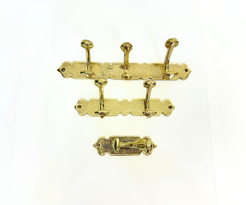 Pode incluir: Conjunto de tr&ecirc;s cabides dourados e um gancho individual. Cada cabide tem uma superf&iacute;cie texturizada e bordas decorativas. Os cabides t&ecirc;m v&aacute;rios ganchos para pendurar itens. O gancho individual tem um design semelhante.