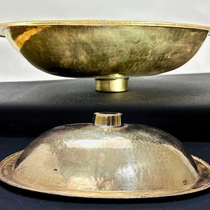 K&ouml;nnte beinhalten: Ein Set aus zwei geh&auml;mmerten Messing-Schalen, eine gr&ouml;&szlig;er als die andere. Die gr&ouml;&szlig;ere Schale hat eine breite, ovale Form und einen erh&ouml;hten Rand. Die kleinere Schale ist ein Deckel f&uuml;r die gr&ouml;&szlig;ere Schale. Beide Schalen haben eine gl&auml;nzende, goldene Oberfl&auml;che.