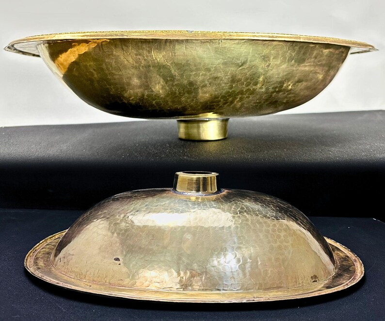 K&ouml;nnte beinhalten: Ein Set aus zwei geh&auml;mmerten Messing-Schalen, eine gr&ouml;&szlig;er als die andere. Die gr&ouml;&szlig;ere Schale hat eine breite, ovale Form und einen erh&ouml;hten Rand. Die kleinere Schale ist ein Deckel f&uuml;r die gr&ouml;&szlig;ere Schale. Beide Schalen haben eine gl&auml;nzende, goldene Oberfl&auml;che.