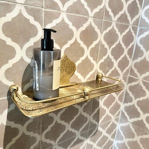 Puede incluir: Estante de ba&ntilde;o de metal dorado con un dise&ntilde;o ornamentado, que sostiene un dispensador de jab&oacute;n gris. El estante est&aacute; montado en una pared alicatada con un patr&oacute;n repetitivo.