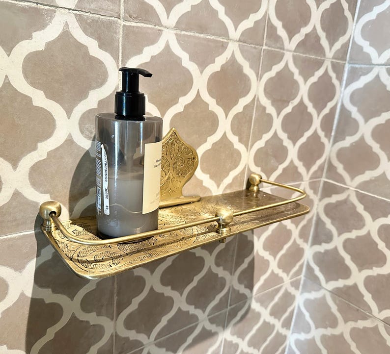 Puede incluir: Estante de ba&ntilde;o de metal dorado con un dise&ntilde;o ornamentado, que sostiene un dispensador de jab&oacute;n gris. El estante est&aacute; montado en una pared alicatada con un patr&oacute;n repetitivo.