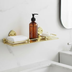 Puede incluir: Estante de ba&ntilde;o dorado con detalles ornamentados, que contiene un dispensador de jab&oacute;n marr&oacute;n, una toalla blanca doblada y una pastilla de jab&oacute;n. El estante est&aacute; montado en una pared de m&aacute;rmol blanco, creando una est&eacute;tica limpia y elegante.