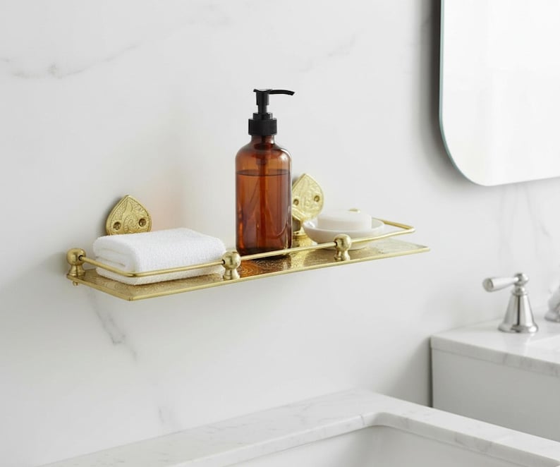 Puede incluir: Estante de ba&ntilde;o dorado con detalles ornamentados, que contiene un dispensador de jab&oacute;n marr&oacute;n, una toalla blanca doblada y una pastilla de jab&oacute;n. El estante est&aacute; montado en una pared de m&aacute;rmol blanco, creando una est&eacute;tica limpia y elegante.