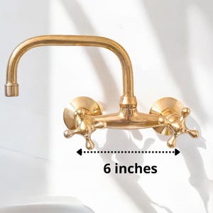 May include: Gold-toned wall-mounted faucet with two handles and a 6-inch spout. The faucet has a traditional design with cross-shaped handles.