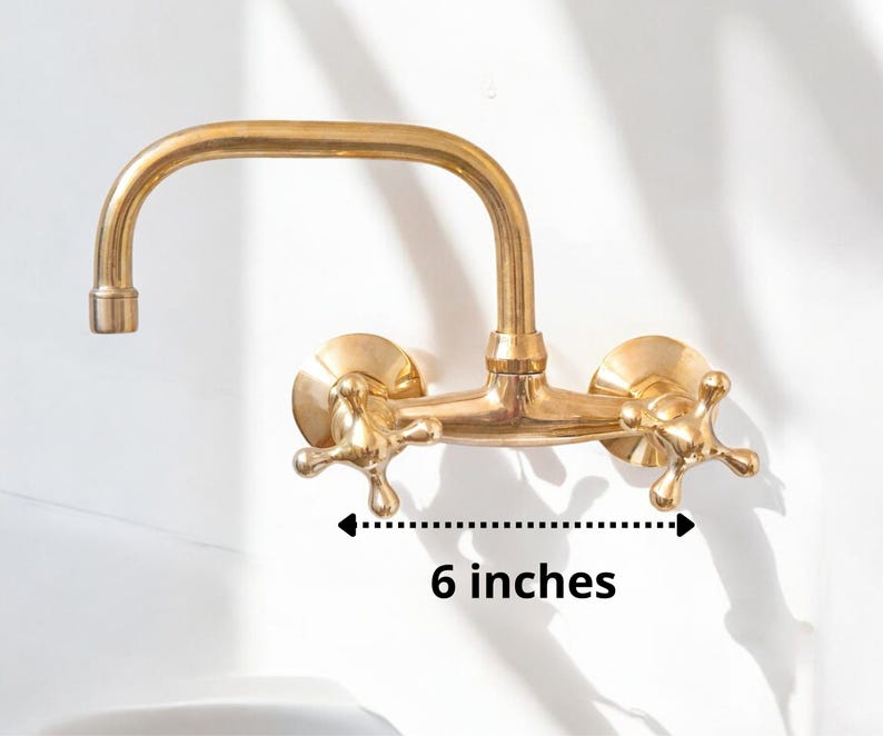 May include: Gold-toned wall-mounted faucet with two handles and a 6-inch spout. The faucet has a traditional design with cross-shaped handles.