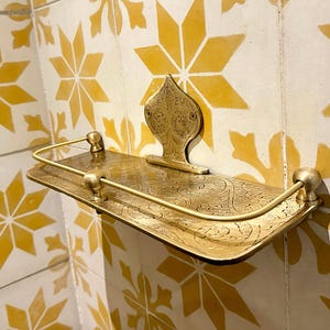 Puede incluir: Estante de metal dorado ornamentado con una placa trasera decorativa y una barandilla curva. El estante tiene un dise&ntilde;o grabado y est&aacute; montado en una pared de azulejos con patrones de estrellas amarillas.