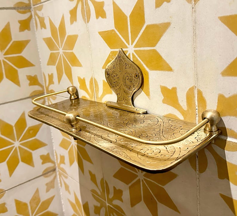 Puede incluir: Estante de metal dorado ornamentado con una placa trasera decorativa y una barandilla curva. El estante tiene un dise&ntilde;o grabado y est&aacute; montado en una pared de azulejos con patrones de estrellas amarillas.