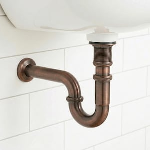 Puede incluir: Un accesorio de fontanería en forma de P de color cobre instalado debajo de un lavabo blanco. La tubería curva está montada en una pared de azulejos blancos. El accesorio tiene una estética vintage o industrial.
