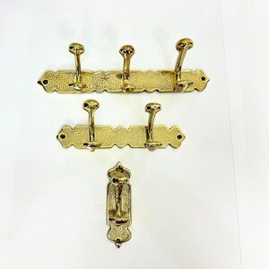 Pode incluir: Tr&ecirc;s cabides de lat&atilde;o com v&aacute;rios ganchos. Os suportes s&atilde;o montados em placas traseiras retangulares texturizadas com bordas decorativas. Os ganchos s&atilde;o curvos e t&ecirc;m um topo arredondado. O acabamento &eacute; dourado brilhante.