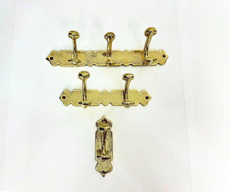 Pode incluir: Tr&ecirc;s cabides de lat&atilde;o com v&aacute;rios ganchos. Os suportes s&atilde;o montados em placas traseiras retangulares texturizadas com bordas decorativas. Os ganchos s&atilde;o curvos e t&ecirc;m um topo arredondado. O acabamento &eacute; dourado brilhante.