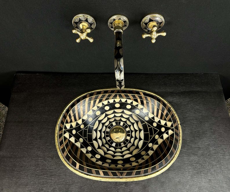 K&ouml;nnte beinhalten: Ein ovales Badezimmerwaschbecken in Schwarz und Gold mit geometrischem Muster. Das Waschbecken hat einen Messinghahn mit zwei Griffen.
