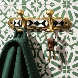 May include: A brass wall-mounted coat rack with three hooks, featuring black and white diamond-shaped accents. A green garment hangs from one hook, and a leather key holder with keys is attached to another. The rack is mounted on a patterned tile wall.