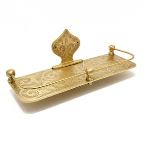 Puede incluir: Un estante de metal dorado con un dise&ntilde;o grabado y ornamentado. El estante tiene una base rectangular con una parte trasera decorativa y una barra curva. El dise&ntilde;o presenta patrones arremolinados y remates decorativos. El estante es probablemente para ba&ntilde;o o decoraci&oacute;n del hogar.
