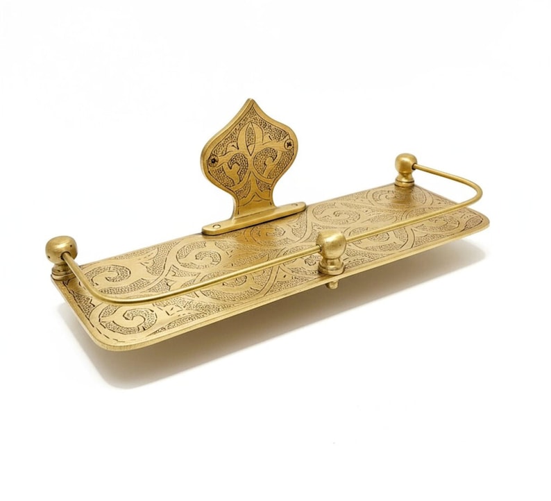 Puede incluir: Un estante de metal dorado con un dise&ntilde;o grabado y ornamentado. El estante tiene una base rectangular con una parte trasera decorativa y una barra curva. El dise&ntilde;o presenta patrones arremolinados y remates decorativos. El estante es probablemente para ba&ntilde;o o decoraci&oacute;n del hogar.