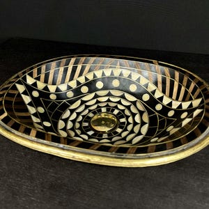 K&ouml;nnte beinhalten: Ovales Waschbecken mit geometrischem Muster in Schwarz und Gold. Das Waschbecken ist aus Holz und hat einen Messingrand.