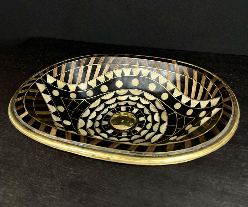 K&ouml;nnte beinhalten: Ovales Waschbecken mit geometrischem Muster in Schwarz und Gold. Das Waschbecken ist aus Holz und hat einen Messingrand.