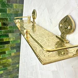 Puede incluir: Estante de metal dorado con un dise&ntilde;o ornamentado y grabado. El estante es rectangular con piezas decorativas en los extremos y una barra curva. El estante est&aacute; montado en una pared de azulejos blancos, con una pared de azulejos verdes en el fondo.