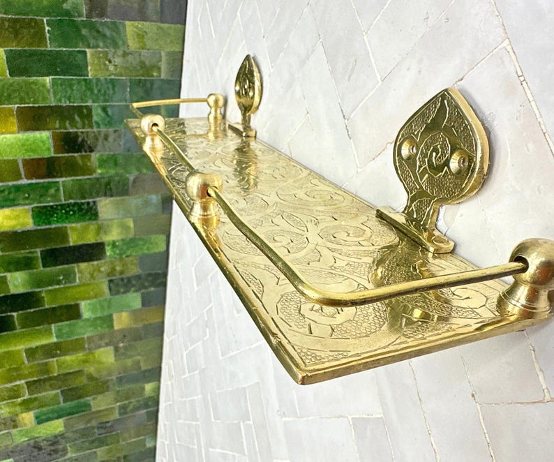 Puede incluir: Estante de metal dorado con un dise&ntilde;o ornamentado y grabado. El estante es rectangular con piezas decorativas en los extremos y una barra curva. El estante est&aacute; montado en una pared de azulejos blancos, con una pared de azulejos verdes en el fondo.