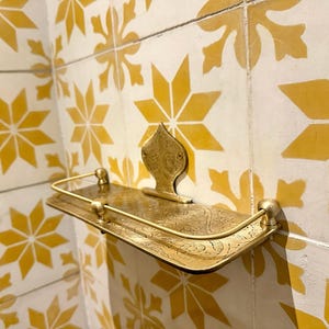 Puede incluir: Estante de metal dorado con un dise&ntilde;o ornamental, montado en una pared con azulejos amarillos y blancos estampados. El estante tiene un frente curvo y una placa trasera decorativa.