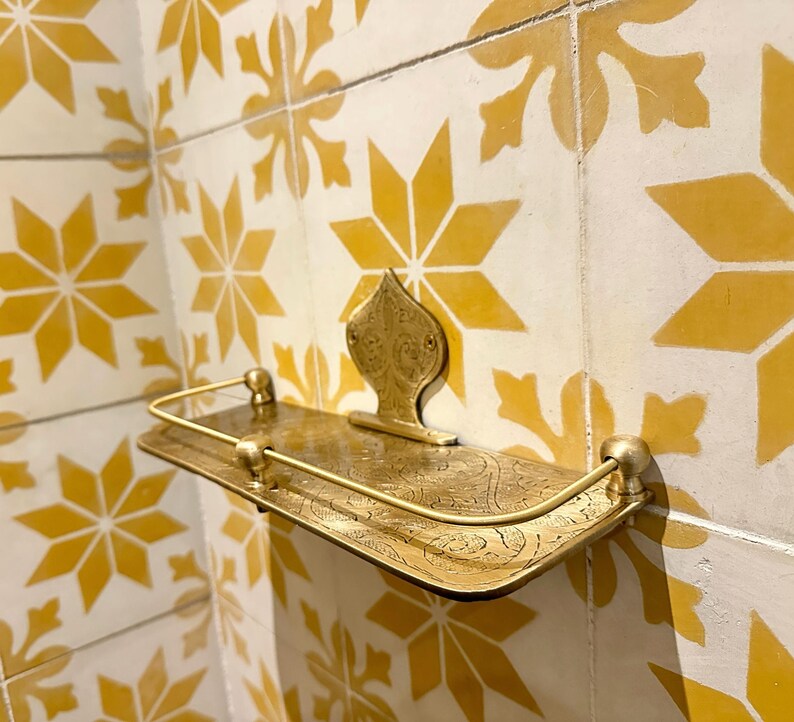 Puede incluir: Estante de metal dorado con un dise&ntilde;o ornamental, montado en una pared con azulejos amarillos y blancos estampados. El estante tiene un frente curvo y una placa trasera decorativa.
