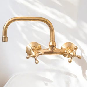 May include: A gold-toned brass wall-mounted faucet with two cross-shaped handles.