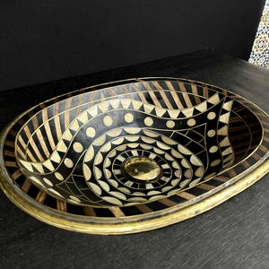 K&ouml;nnte beinhalten: Ovales Waschbecken mit geometrischem Muster in Schwarz und Wei&szlig;. Das Waschbecken ist aus Holz und hat einen Messingrand.