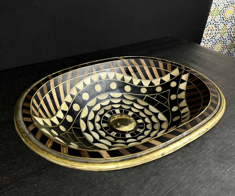 K&ouml;nnte beinhalten: Ovales Waschbecken mit geometrischem Muster in Schwarz und Wei&szlig;. Das Waschbecken ist aus Holz und hat einen Messingrand.
