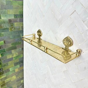 Puede incluir: Un estante de metal dorado con patrones florales decorativos y piezas finales ornamentadas. El estante est&aacute; montado en una pared de azulejos blancos, con una pared de azulejos verdes visible en el lado izquierdo de la imagen.