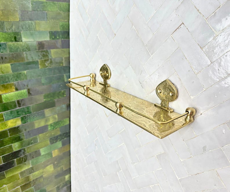 Puede incluir: Un estante de metal dorado con patrones florales decorativos y piezas finales ornamentadas. El estante est&aacute; montado en una pared de azulejos blancos, con una pared de azulejos verdes visible en el lado izquierdo de la imagen.