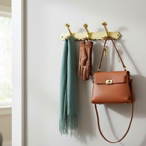 Pode incluir: Um cabide de parede de lat&atilde;o com tr&ecirc;s ganchos segura um len&ccedil;o verde-azulado, luvas de couro marrom e uma bolsa de couro marrom. A bolsa tem um fecho dourado e uma longa al&ccedil;a de ombro. A parede &eacute; pintada de cinza claro.