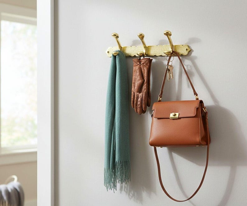 Pode incluir: Um cabide de parede de lat&atilde;o com tr&ecirc;s ganchos segura um len&ccedil;o verde-azulado, luvas de couro marrom e uma bolsa de couro marrom. A bolsa tem um fecho dourado e uma longa al&ccedil;a de ombro. A parede &eacute; pintada de cinza claro.