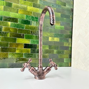 Può includere: Un rubinetto color rame con una texture martellata. Il rubinetto ha un beccuccio curvo e due maniglie a forma di croce. Lo sfondo presenta una parete di piastrelle verdi.