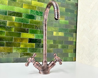 May include: A copper-coloured faucet with a hammered texture. The faucet has a curved spout and two cross-shaped handles. The background features a green tile wall.