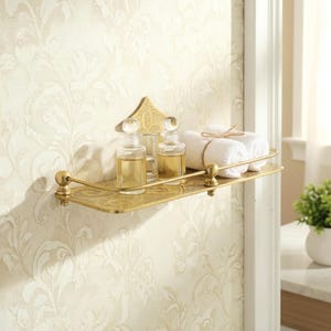 Puede incluir: Estante de ba&ntilde;o dorado y ornamentado con una placa trasera decorativa. El estante contiene dos botellas de vidrio con tapones transparentes, un vaso y dos toallas blancas enrolladas atadas con hilo. El estante est&aacute; montado en una pared con papel pintado floral.