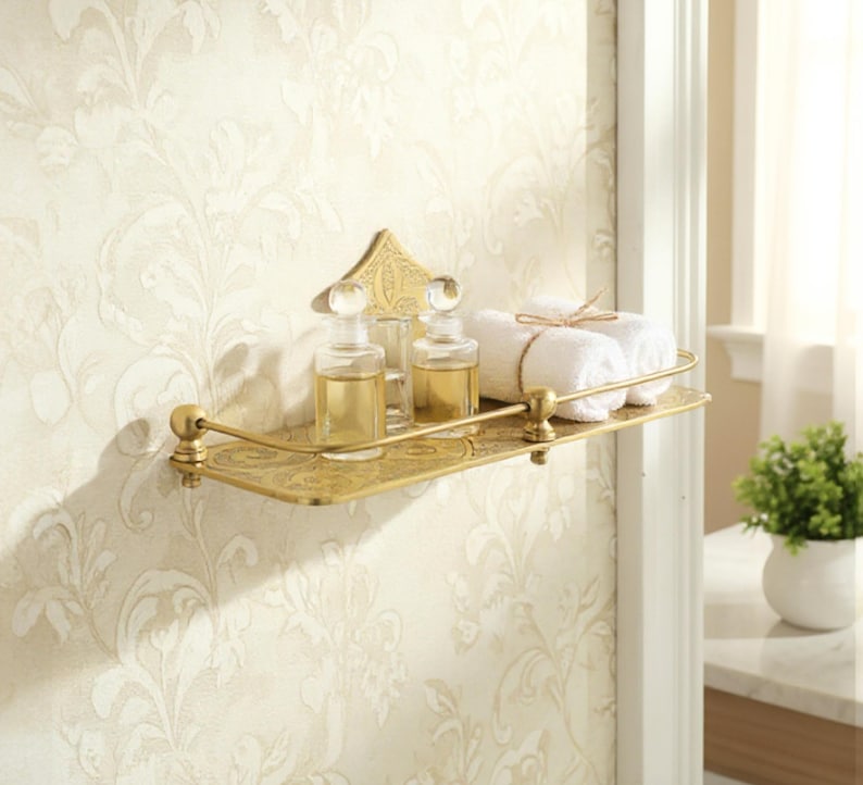 Puede incluir: Estante de ba&ntilde;o dorado y ornamentado con una placa trasera decorativa. El estante contiene dos botellas de vidrio con tapones transparentes, un vaso y dos toallas blancas enrolladas atadas con hilo. El estante est&aacute; montado en una pared con papel pintado floral.