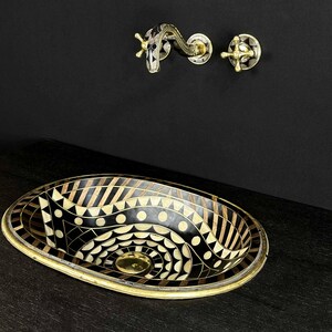 K&ouml;nnte beinhalten: Ein ovales Waschbecken in Schwarz und Gold mit einem geometrischen Muster. Das Waschbecken besteht aus einer Mosaik aus verschiedenen Materialien, darunter Holz, Metall und Stein. Das Waschbecken steht auf einer schwarzen Arbeitsplatte.