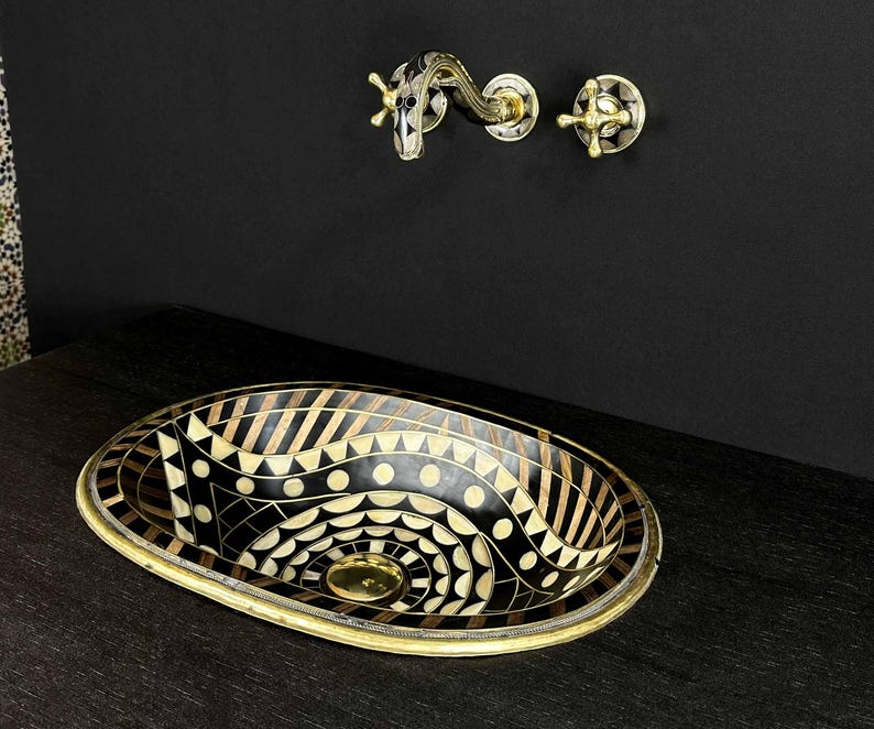 K&ouml;nnte beinhalten: Ein ovales Waschbecken in Schwarz und Gold mit einem geometrischen Muster. Das Waschbecken besteht aus einer Mosaik aus verschiedenen Materialien, darunter Holz, Metall und Stein. Das Waschbecken steht auf einer schwarzen Arbeitsplatte.