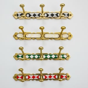 Pode incluir: Quatro cabides decorativos de latão, cada um com três ganchos. Cada cabide apresenta um padrão diferente: losangos em preto e branco, retângulos brancos, losangos em verde e branco e losangos em vermelho e branco. Os ganchos têm um design arredondado.