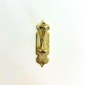 Pode incluir: Um gancho de parede dourado e ornamentado com um design decorativo. O gancho tem uma superf&iacute;cie texturizada e uma parte superior e inferior arredondadas. O gancho &eacute; montado numa placa traseira retangular com um padr&atilde;o detalhado. O design geral &eacute; vintage e elegante.