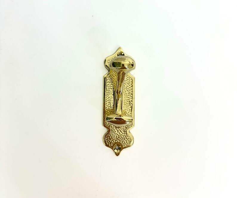 Pode incluir: Um gancho de parede dourado e ornamentado com um design decorativo. O gancho tem uma superf&iacute;cie texturizada e uma parte superior e inferior arredondadas. O gancho &eacute; montado numa placa traseira retangular com um padr&atilde;o detalhado. O design geral &eacute; vintage e elegante.
