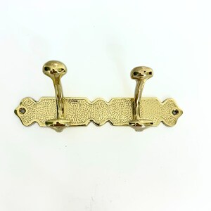 Pode incluir: Um suporte de parede com gancho duplo dourado. Os ganchos s&atilde;o curvos com pontas arredondadas, presos a uma placa traseira retangular texturizada com bordas decorativas. A placa traseira tem um padr&atilde;o pontilhado.