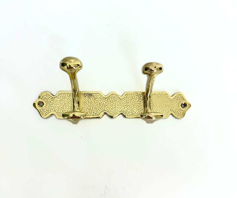 Pode incluir: Um suporte de parede com gancho duplo dourado. Os ganchos s&atilde;o curvos com pontas arredondadas, presos a uma placa traseira retangular texturizada com bordas decorativas. A placa traseira tem um padr&atilde;o pontilhado.