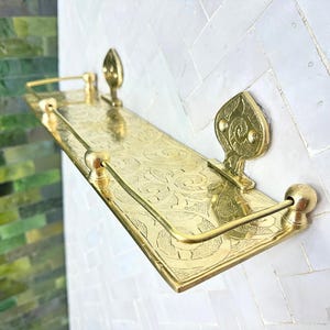 Puede incluir: Estante rectangular de lat&oacute;n con un patr&oacute;n floral adornado y piezas decorativas en los extremos. El estante tiene un borde elevado y est&aacute; montado en una pared de azulejos blancos. El estante mide aproximadamente 30 cm de largo.