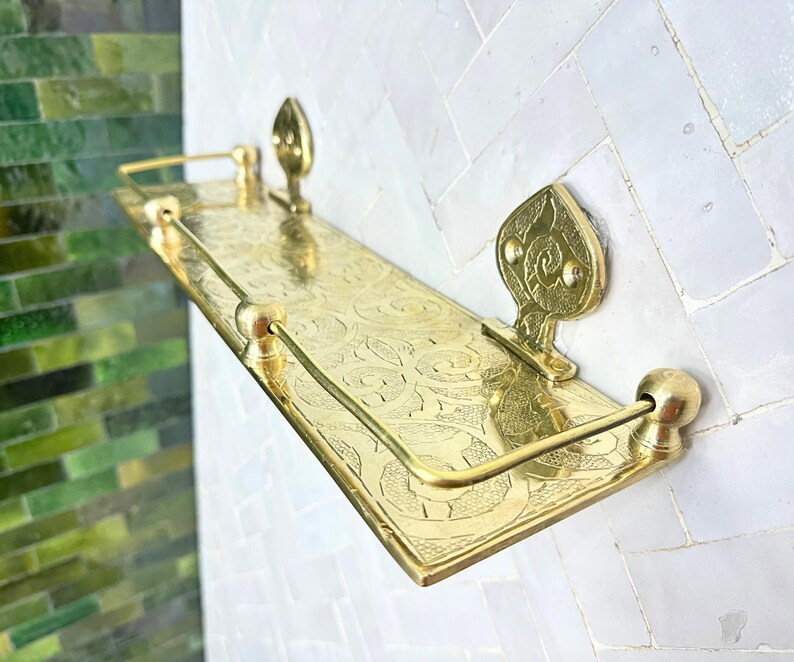 Puede incluir: Estante rectangular de lat&oacute;n con un patr&oacute;n floral adornado y piezas decorativas en los extremos. El estante tiene un borde elevado y est&aacute; montado en una pared de azulejos blancos. El estante mide aproximadamente 30 cm de largo.