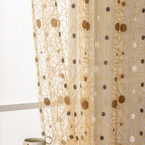 Peut inclure: Rideau transparent de couleur claire avec un motif doré tourbillonnant et des cercles marron, gris et blancs épars. Le rideau est suspendu devant une fenêtre. Une tasse et une soucoupe à motifs floraux sont posées sur une table.
