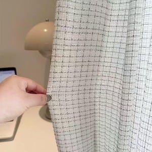 Puede incluir: Primer plano de una tela de color claro con un patrón de cuadrícula de líneas negras. La tela parece ser una cortina o un textil similar. La textura es ligeramente áspera y el aspecto general es neutro y versátil.