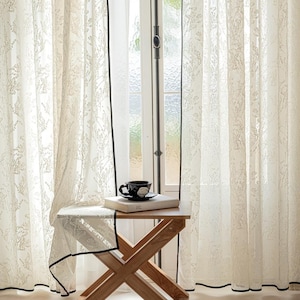 May include: Delicate white floral patterned curtains with black edging frame a window. A small wooden table holds a book, a cup, and a saucer. The curtains are partially drawn, allowing natural light to filter through.