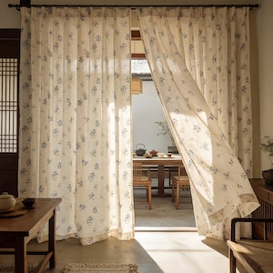 May include: Floor-length curtains made of off-white fabric featuring a repeating pattern of small blue floral sprigs. The curtains are hung on a dark rod and are partially parted, revealing a view of a wooden table and chairs in a bright room.
